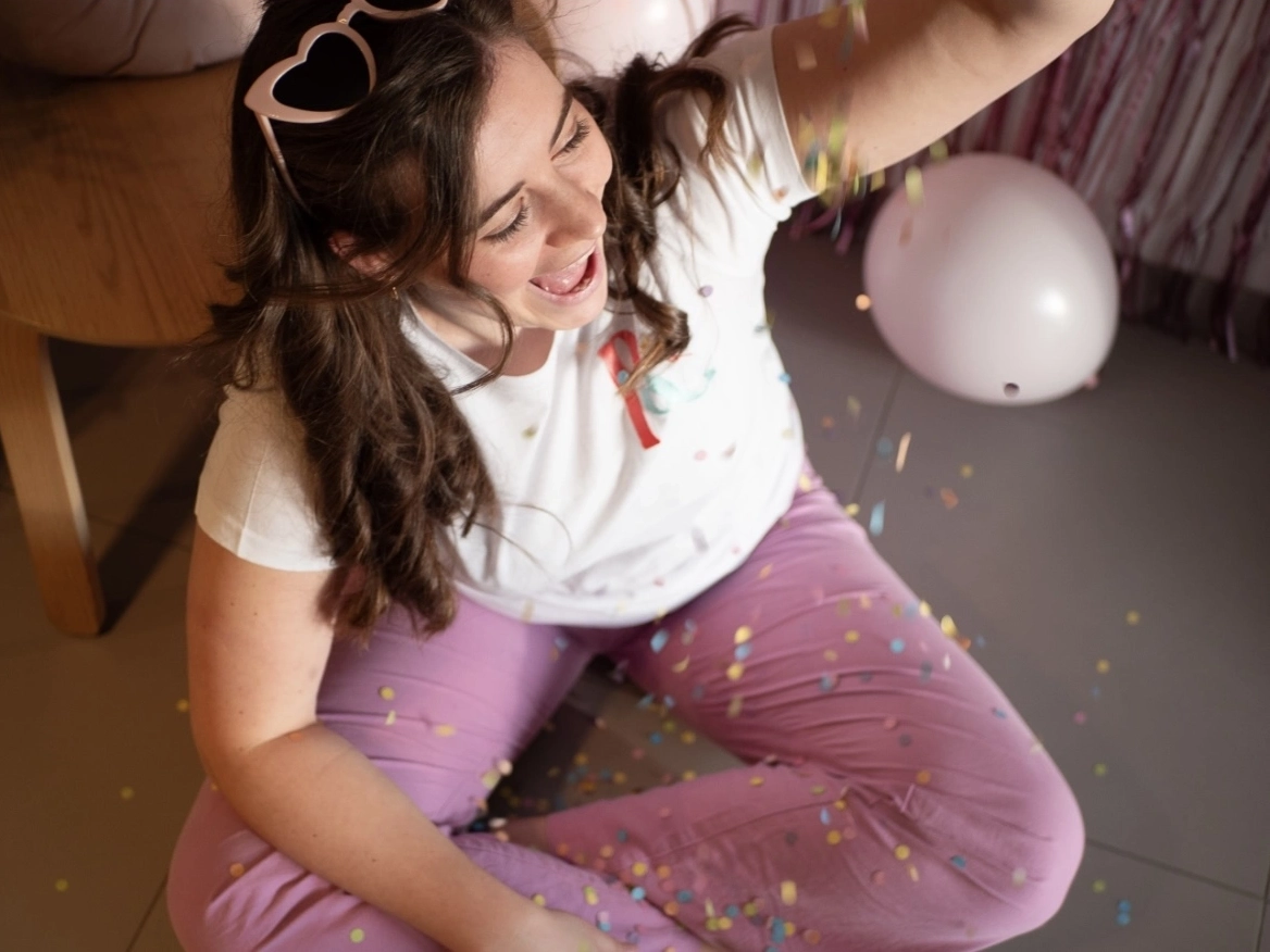 Vrouw gooit confetti in de lucht tijdens een feest, zittend op de vloer in een paarse broek.