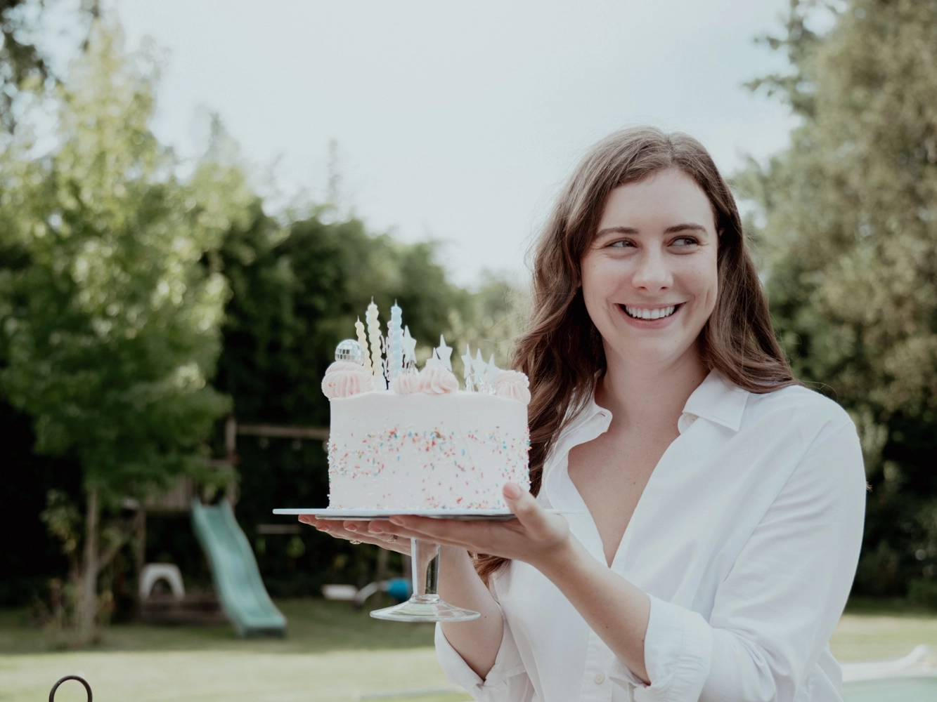 Vrouw toont roze verjaardagstaart in een tuin met zwembad.
